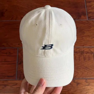 A hand holding up a cream baseball cap with a single dark letter embroidered on the front panel