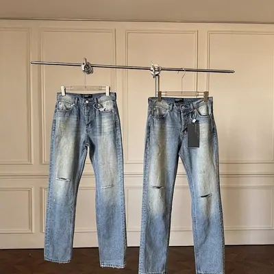 Two pairs of light-wash jeans hanging on a rail in front of a panelled wall, the right-hand pair torn open at the knee