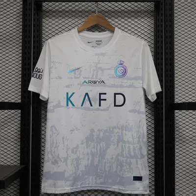 A white short-sleeve football shirt on a wooden hanger against a dark mesh panel, with lettering across the chest and a small crest above it