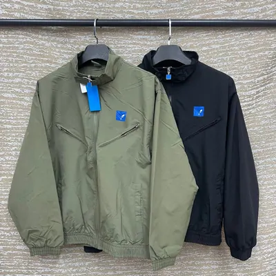 Two lightweight zip-front jackets on hangers against a grey drape, one sage green and one navy, each with a small blue tag at the chest