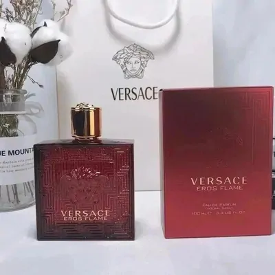 A dark red fragrance bottle standing beside its matching red carton and a printed paper bag, on a pale surface