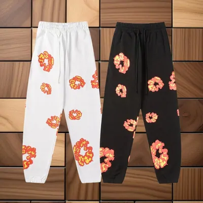 Two pairs of cuffed sweatpants laid on a wooden floor, one cream and one black, both printed all over with orange flower heads