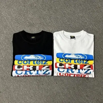 Two short-sleeve tees folded side by side on a grey mat, one black and one white, both carrying the same blocky red and blue chest print